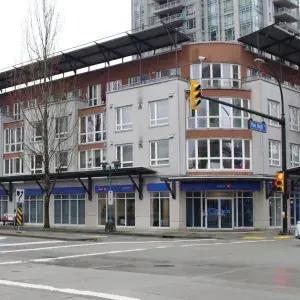 Exterior photo of 1-1161 The High Street with a modern multi-story building featuring large windows and a bank at street level. Traffic lights and bare trees line the road.