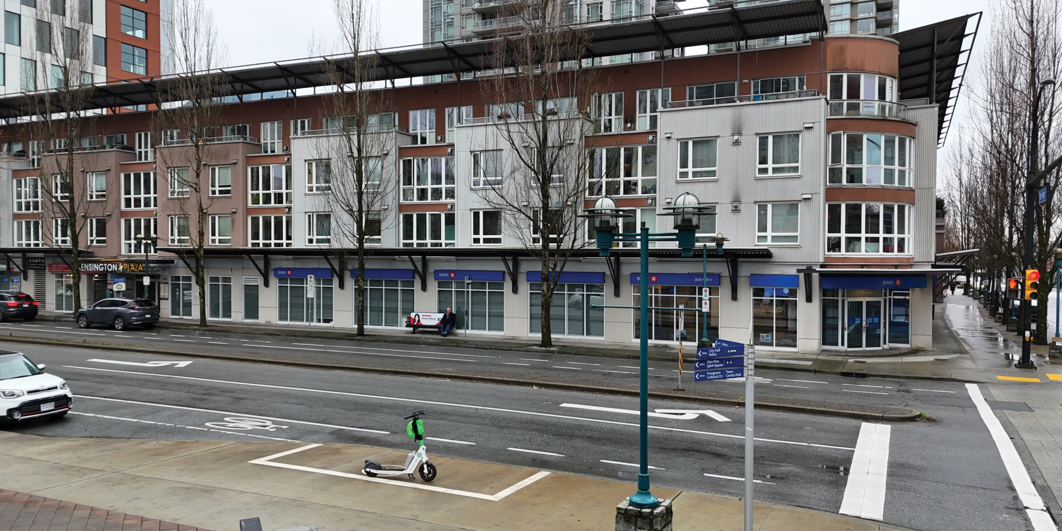 Exterior photo of 1-1161 The High Street with a modern multi-story building featuring large windows and a bank at street level. Traffic lights and bare trees line the road.