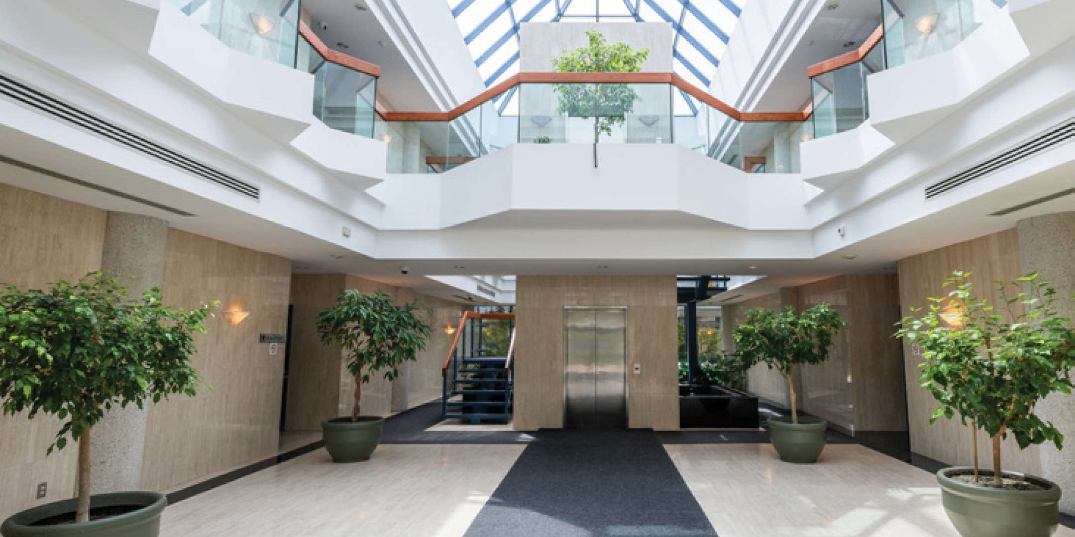 Ground floor view of Atrium of 2 storey office building with cream walls, skylight and several indoor trees as accents