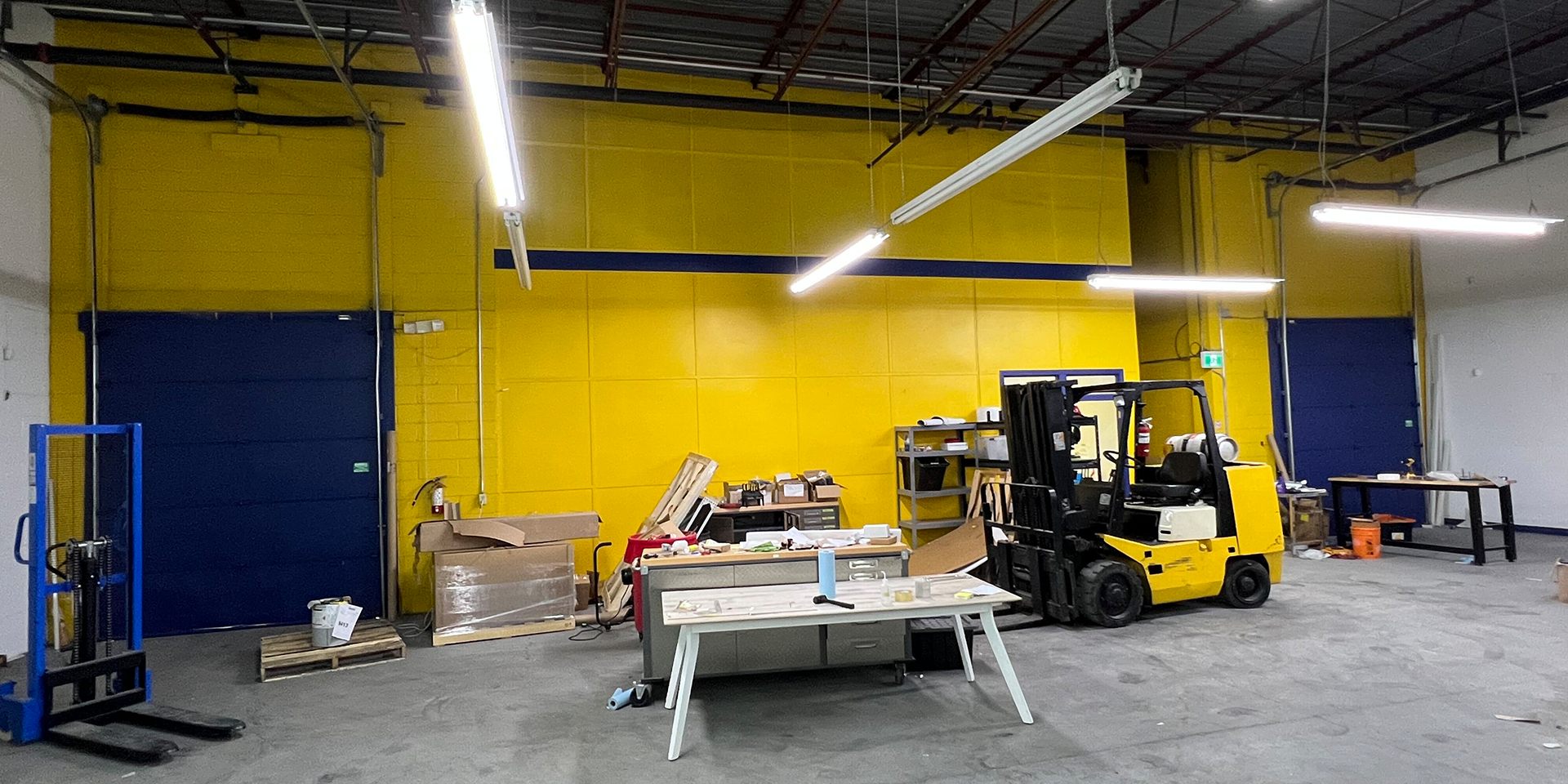455 Cochrane Drive 24-25 another angle of the warehouse showing forklift and workbenches sit before a yellow wall with blue doors.