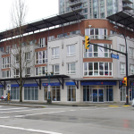 Exterior photo of 1-1161 The High Street with a modern multi-story building featuring large windows and a bank at street level. Traffic lights and bare trees line the road.