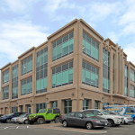 three story office building with cars parked in front. reddish brock with green glass